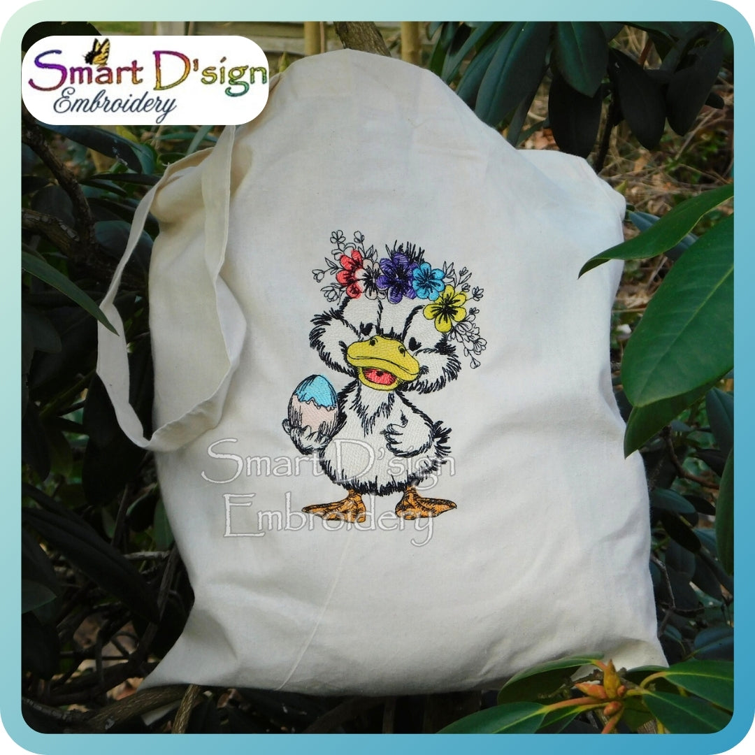 White tote bag with embroidered design of a duck with flowers on its head, placed outdoors.